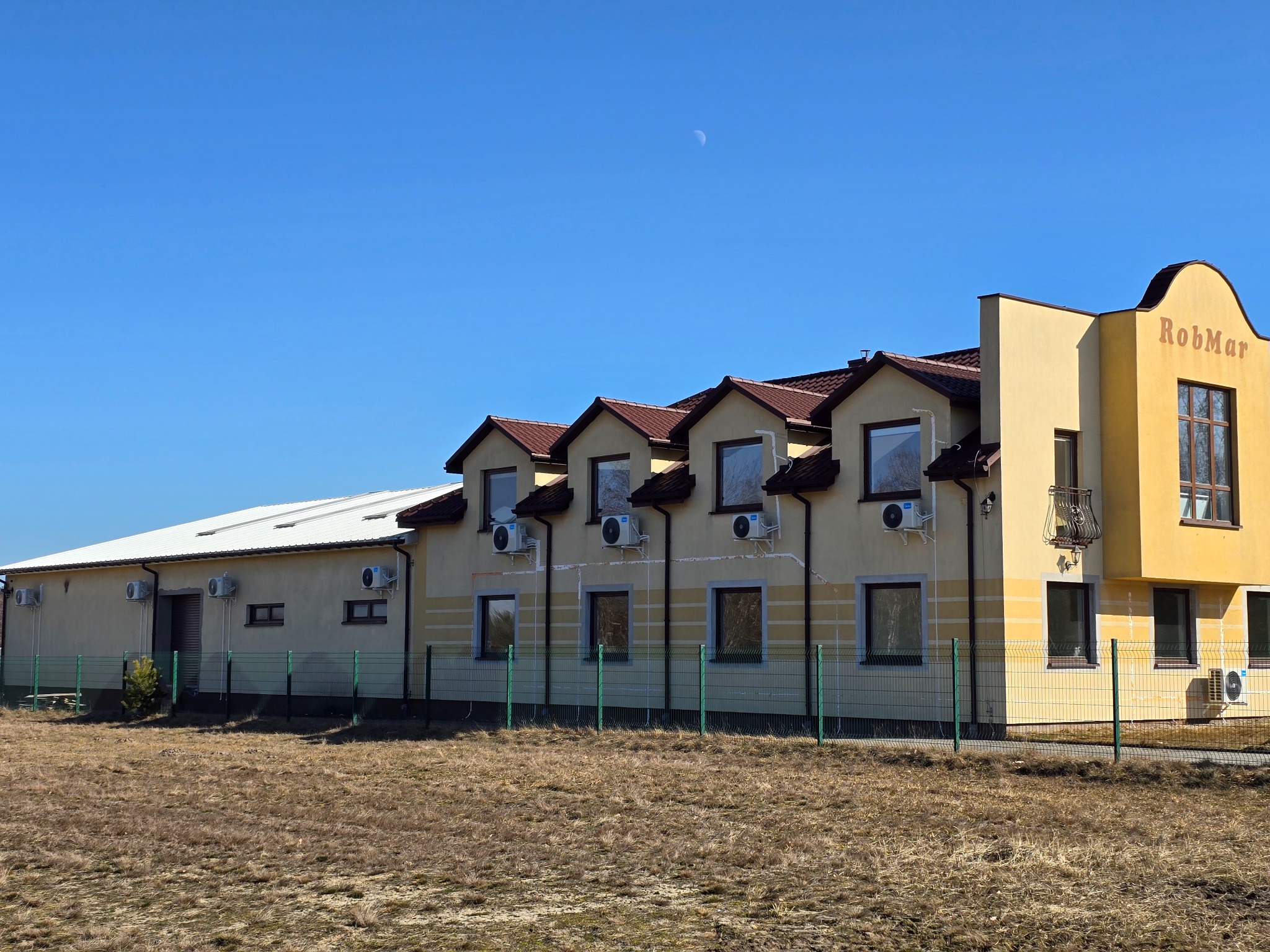 Widok z zewnątrz budynku z zainstalowanymi klimatyzatorami na ścianach, widoczny zielony płot i błękitne niebo z księżycem w tle.