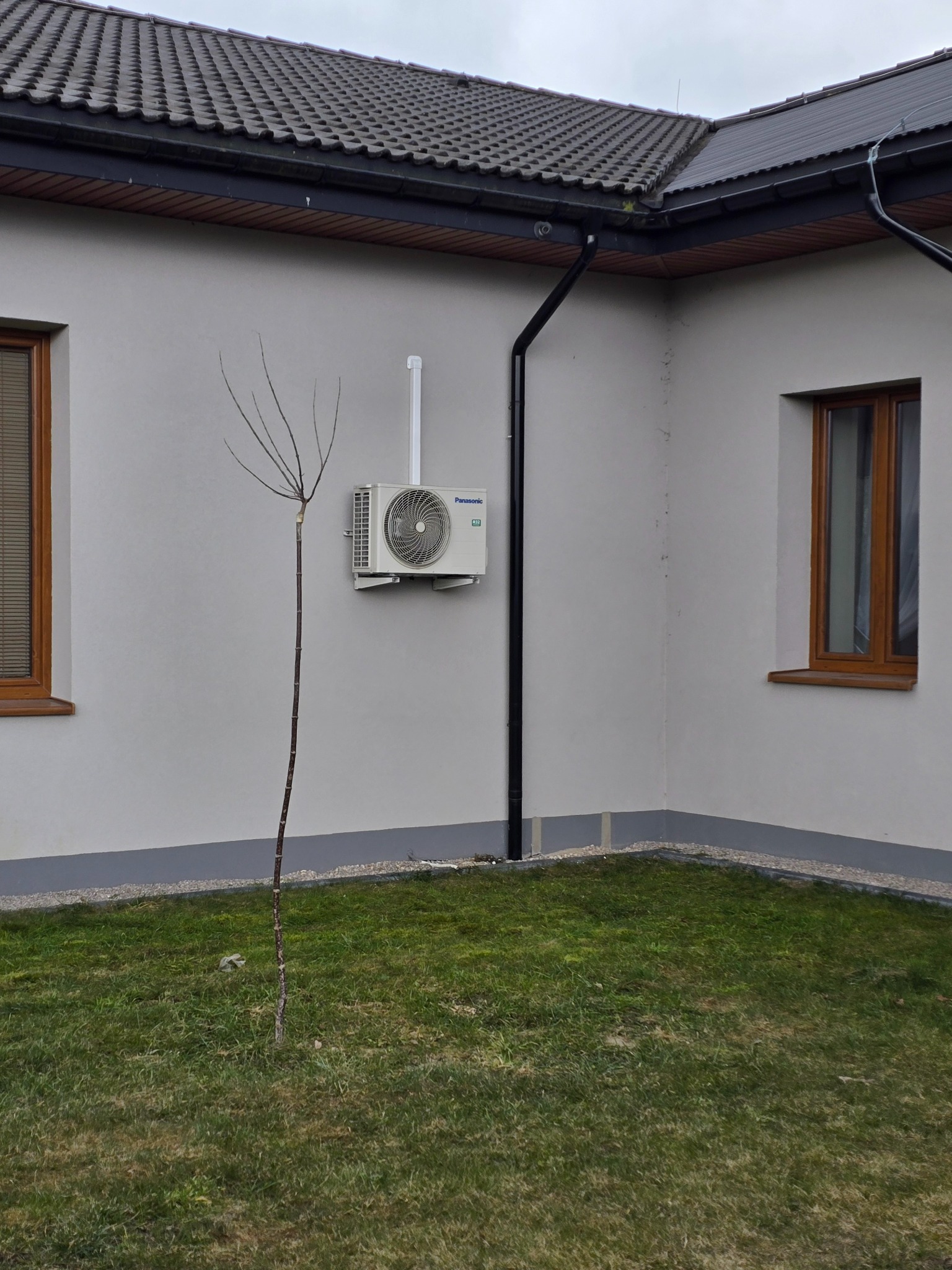 Zewnętrzna jednostka klimatyzatora Panasonic zamontowana na elewacji budynku we Włoszczowie, obok młodego drzewka bez liści i okna z brązową ramą.