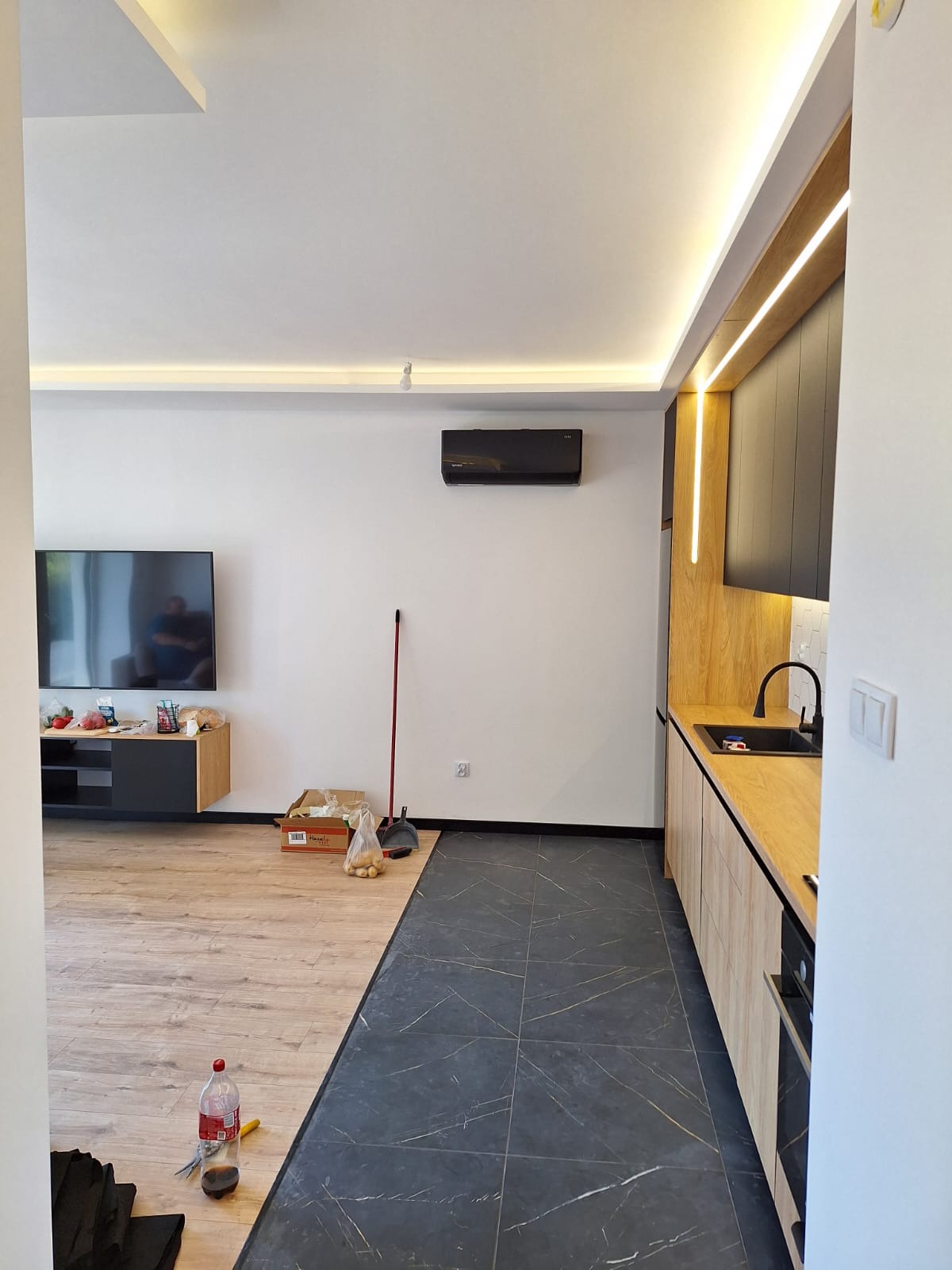 Czarny klimatyzator ścienny w nowoczesnym salonie z aneksem kuchennym, widoczne kartony, mop i inne przedmioty sugerujące trwający remont.