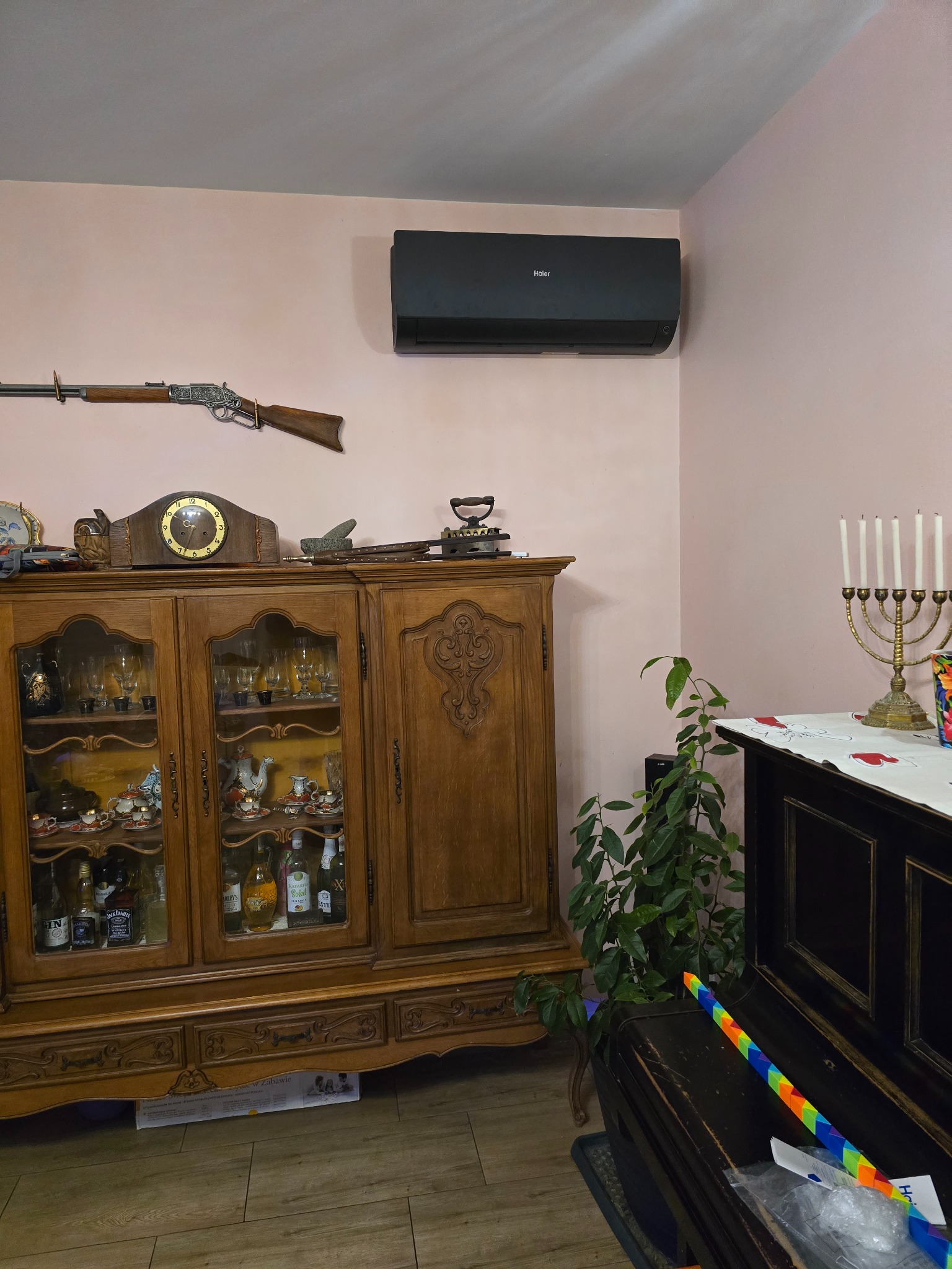 Czarny klimatyzator marki Haier zamontowany na różowej ścianie w pokoju z antykami: karabin na ścianie, stary kredens z porcelaną i alkoholem, świecznik chanukowy na fortepianie.