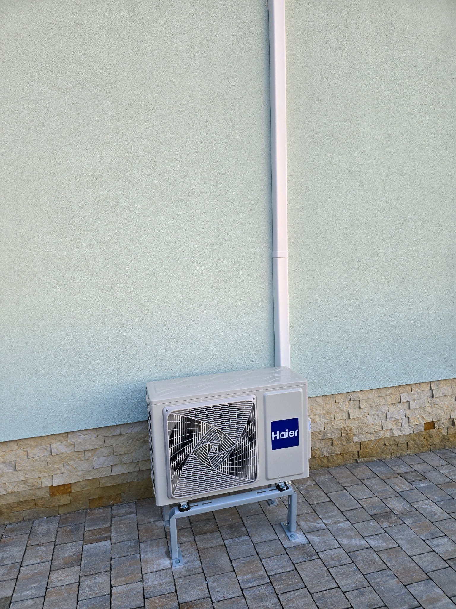 Zewnętrzna jednostka klimatyzacji Haier zamontowana na metalowym stelażu przy jasnej elewacji budynku z dekoracyjnym cokołem z kamienia.