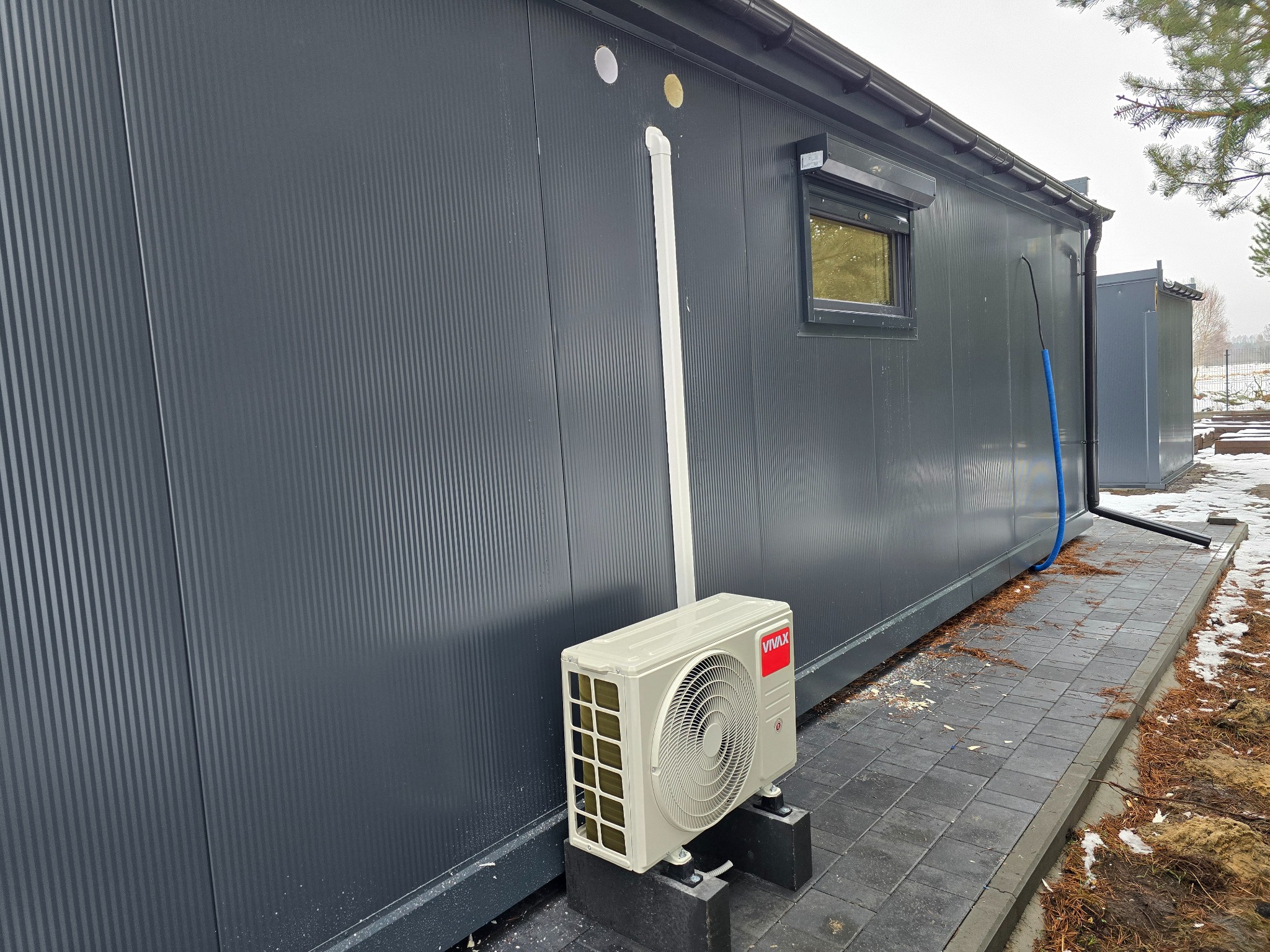 Zewnętrzna jednostka klimatyzacji VIVAX zamontowana na betonowych podstawach przy szarej, modułowej ścianie budynku z widocznymi rurami instalacyjnymi i śladami montażu na kostce brukowej.