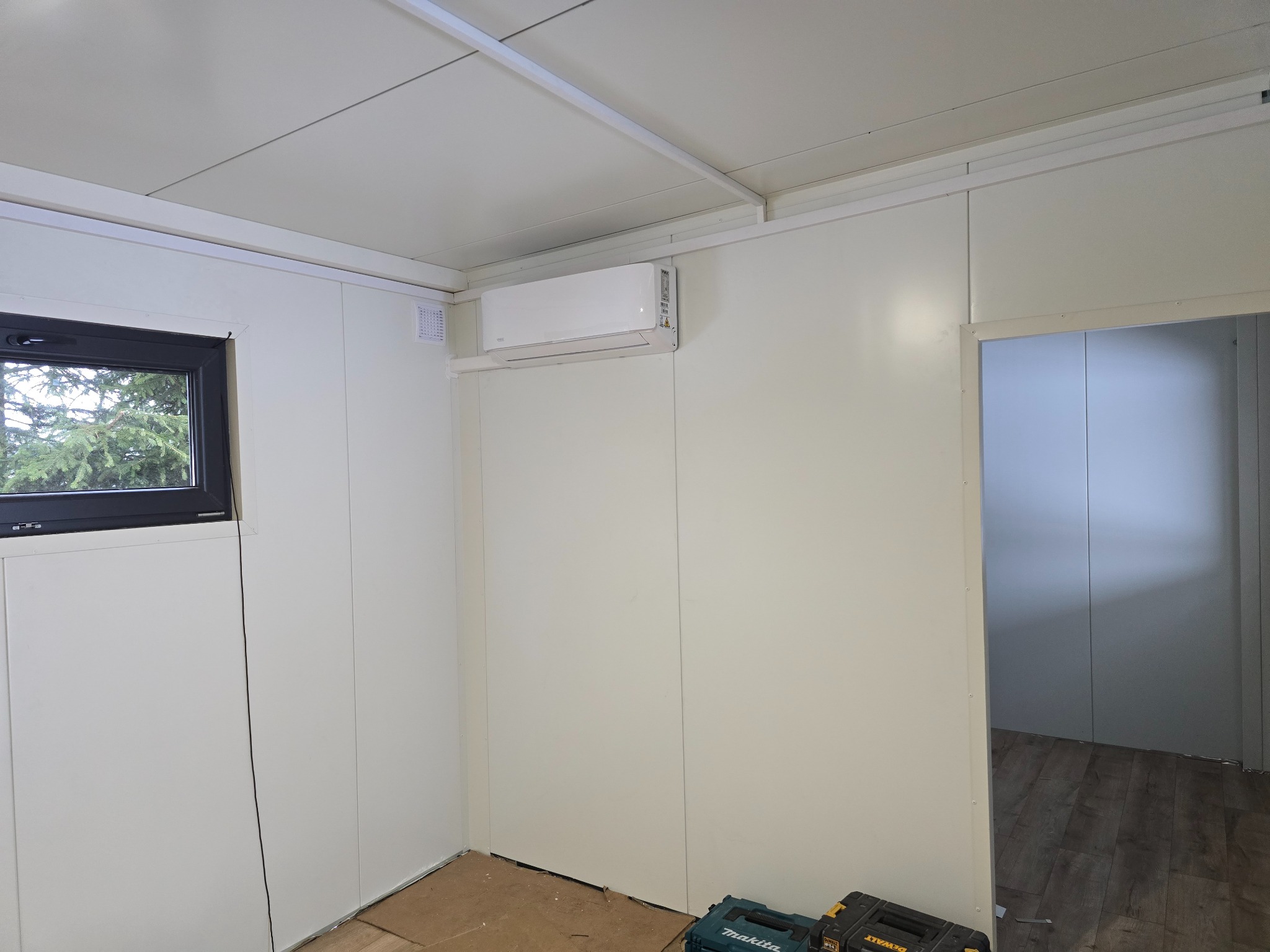 Wnętrze pomieszczenia z zamontowanym, białym klimatyzatorem ściennym, widoczne narzędzia w skrzynkach na podłodze i okno z widokiem na zieleń.