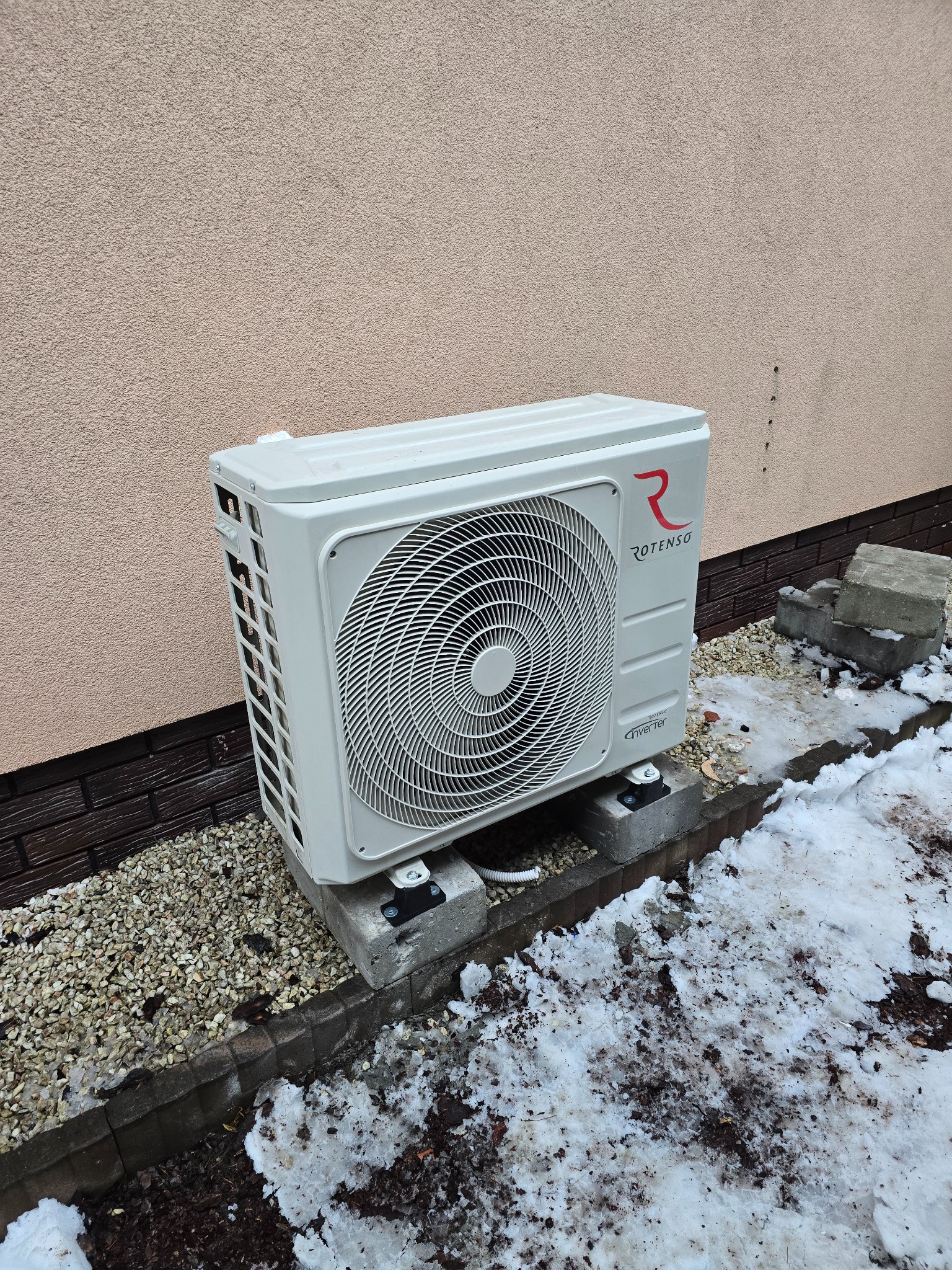 Zewnętrzna jednostka klimatyzatora Rotenso Inverter zamontowana na betonowych bloczkach przy ścianie budynku, widoczne resztki śniegu.