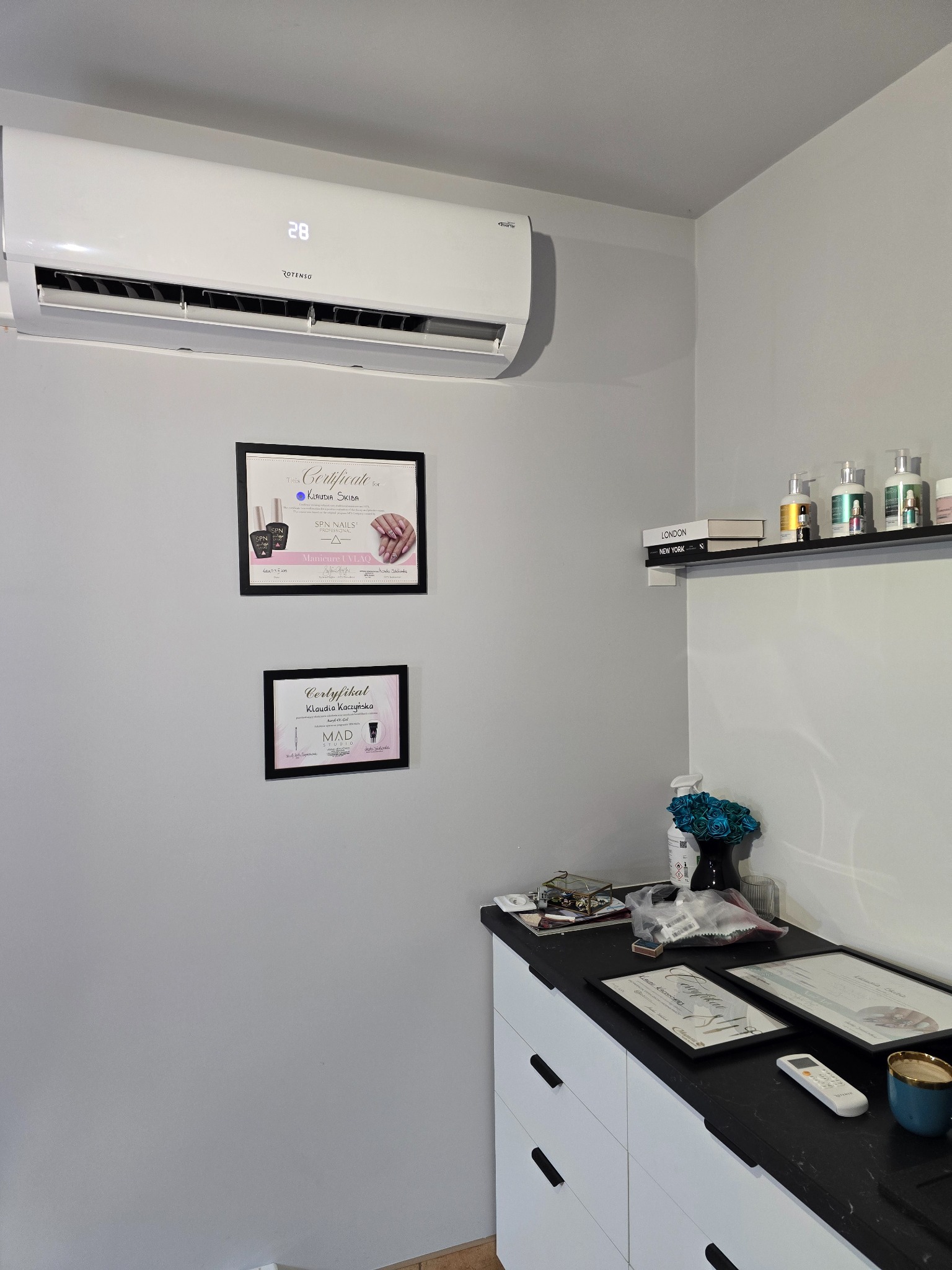 Biały klimatyzator marki Rotenso zamontowany na szarej ścianie w salonie kosmetycznym, pod nim wiszą dwa certyfikaty w czarnych ramkach, a obok na półce stoją książki i kosmetyki.