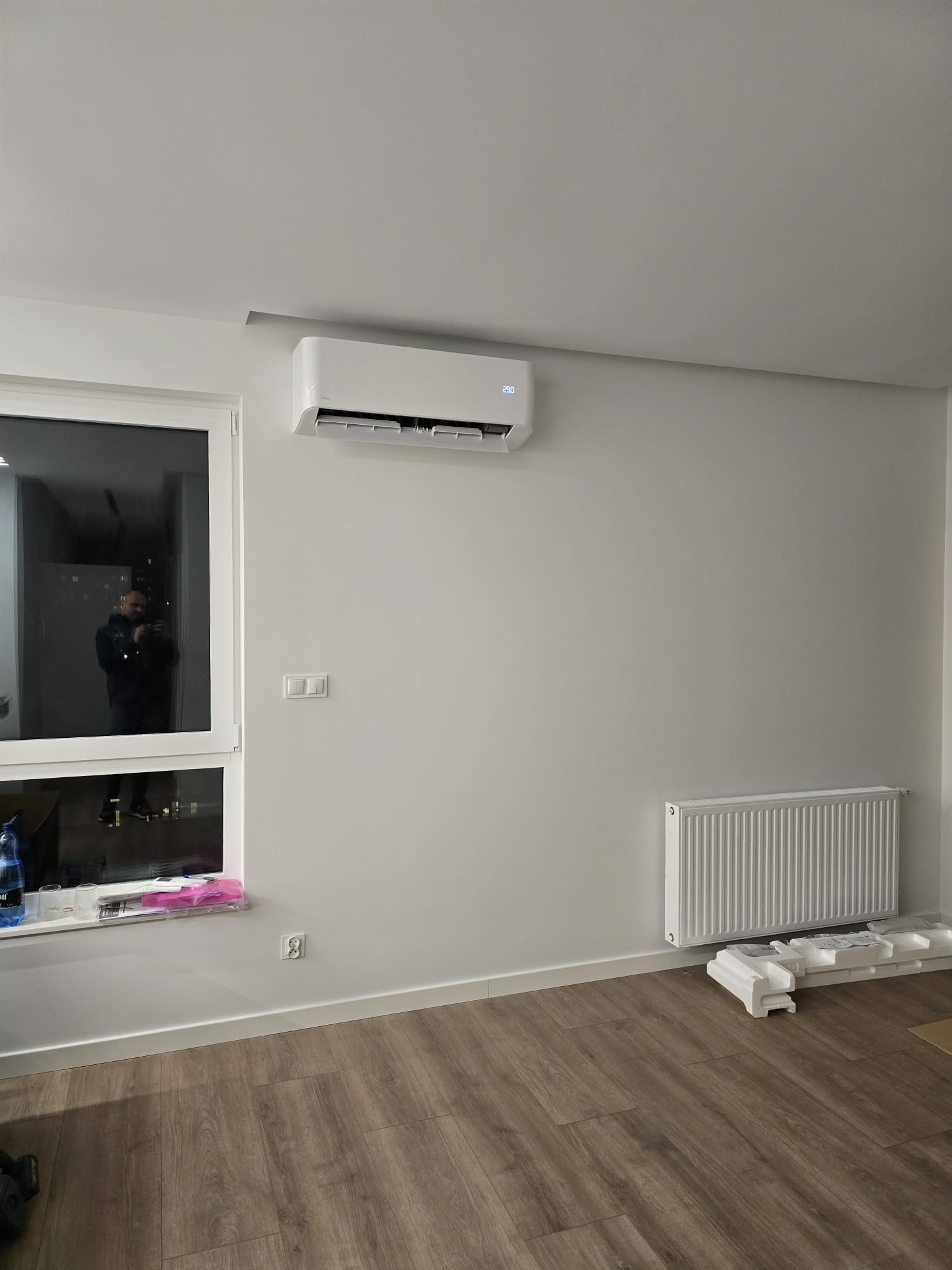 Biały klimatyzator ścienny z wyświetlaczem ustawionej temperatury, zamontowany w nowoczesnym wnętrzu z drewnianą podłogą, białą ścianą, grzejnikiem i oknem z widokiem na miasto nocą.