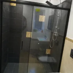 Nowoczesna kabina prysznicowa z czarną ramą i przesuwanymi drzwiami, wykończona ciemnymi płytkami z dekoracyjnymi, złotymi akcentami na ścianach, widok na toaletę i umywalkę w tle.