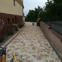 Ułożona kostka brukowa wzdłuż budynku z narzędziami i zagęszczarką w rogu, widok na w&oacute;zek i zieleń w tle.