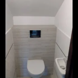 Wąska toaleta z umywalką, wykończona w jasnych barwach z akcentem drewnopodobnych płytek na ścianie za muszlą klozetową i minimalistycznym panelem spłukującym.