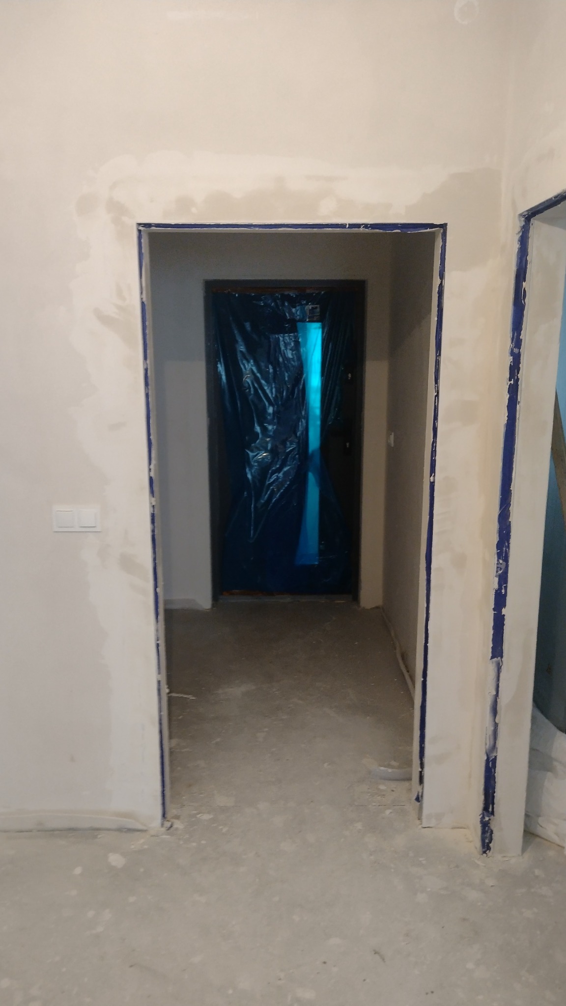 Przygotowanie do montażu ościeżnic w trakcie prac wykończeniowych: ściany po gruntowaniu, zabezpieczone taśmą malarską otwory drzwiowe, drzwi wejściowe osłonięte folią.