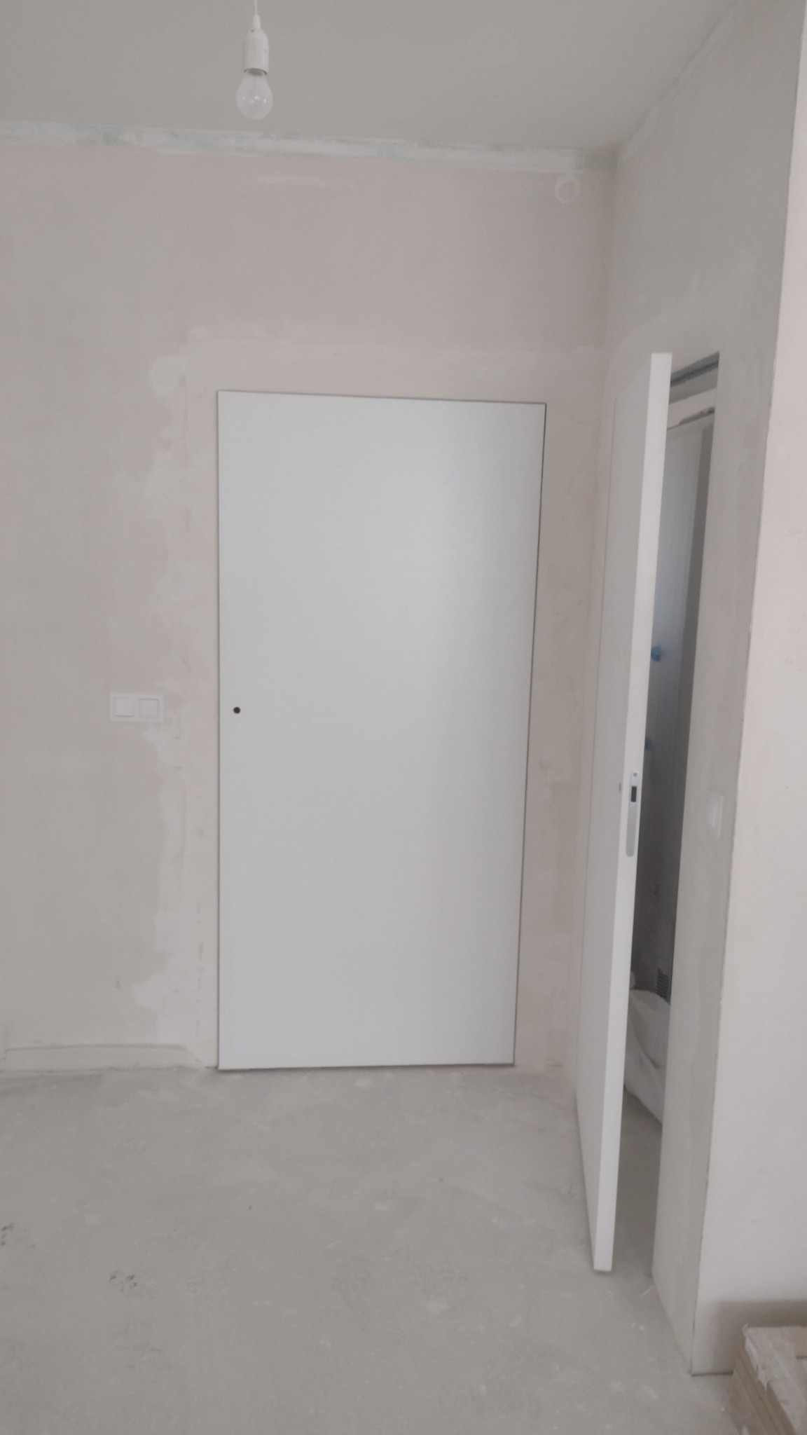 Białe, minimalistyczne drzwi wewnętrzne, częściowo otwarte, widoczne na tle surowych ścian w trakcie wykończenia; sufit z wiszącą żarówką.