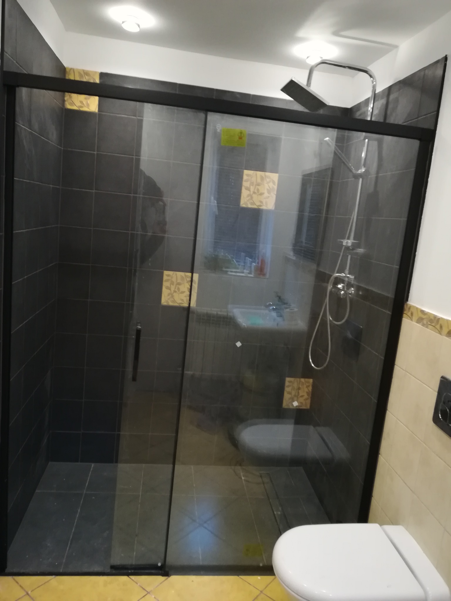 Nowoczesna kabina prysznicowa z czarną ramą i przesuwanymi drzwiami, wykończona ciemnymi płytkami z dekoracyjnymi, złotymi akcentami na ścianach, widok na toaletę i umywalkę w tle.