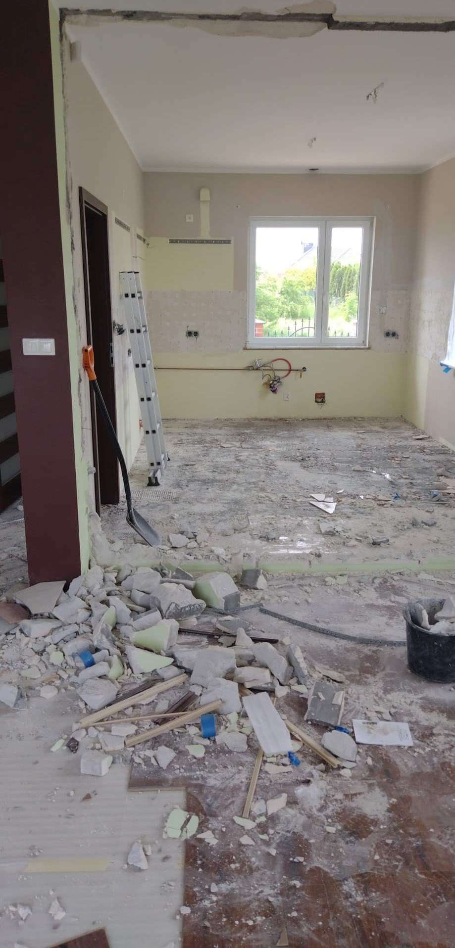 Demontaż płytek w kuchni podczas remontu: widoczny bałagan, gruz, odłupane fragmenty płytek na podłodze, drabina oparta o ścianę, łopata i wiadro w tle