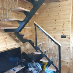 Drewniane schody z czarną, metalową konstrukcją i linową balustradą w drewnianym wnętrzu. U dołu widoczne narzędzia, w tym piła elektryczna, sugerujące trwający montaż.