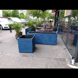 Niebieskie, drewniane donice na zewnątrz lokalu gastronomicznego, z widocznymi roślinami w tle i fragmentem chodnika z kostki.