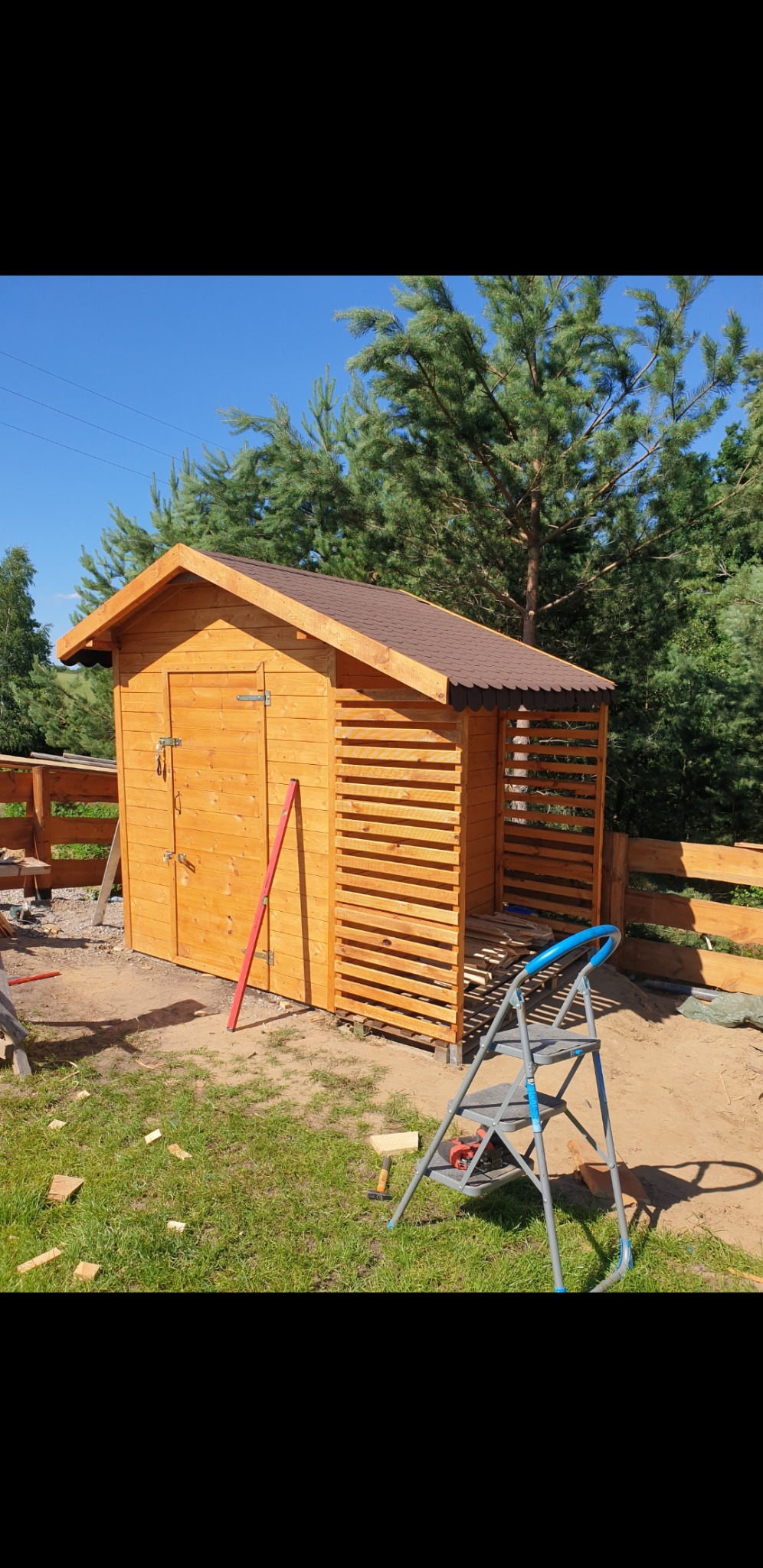 Drewniana altana ogrodowa z brązowym dachem, widoczna w trakcie budowy, z poziomnicą opartą o ścianę i drabinką obok, otoczona zielenią i drzewami.