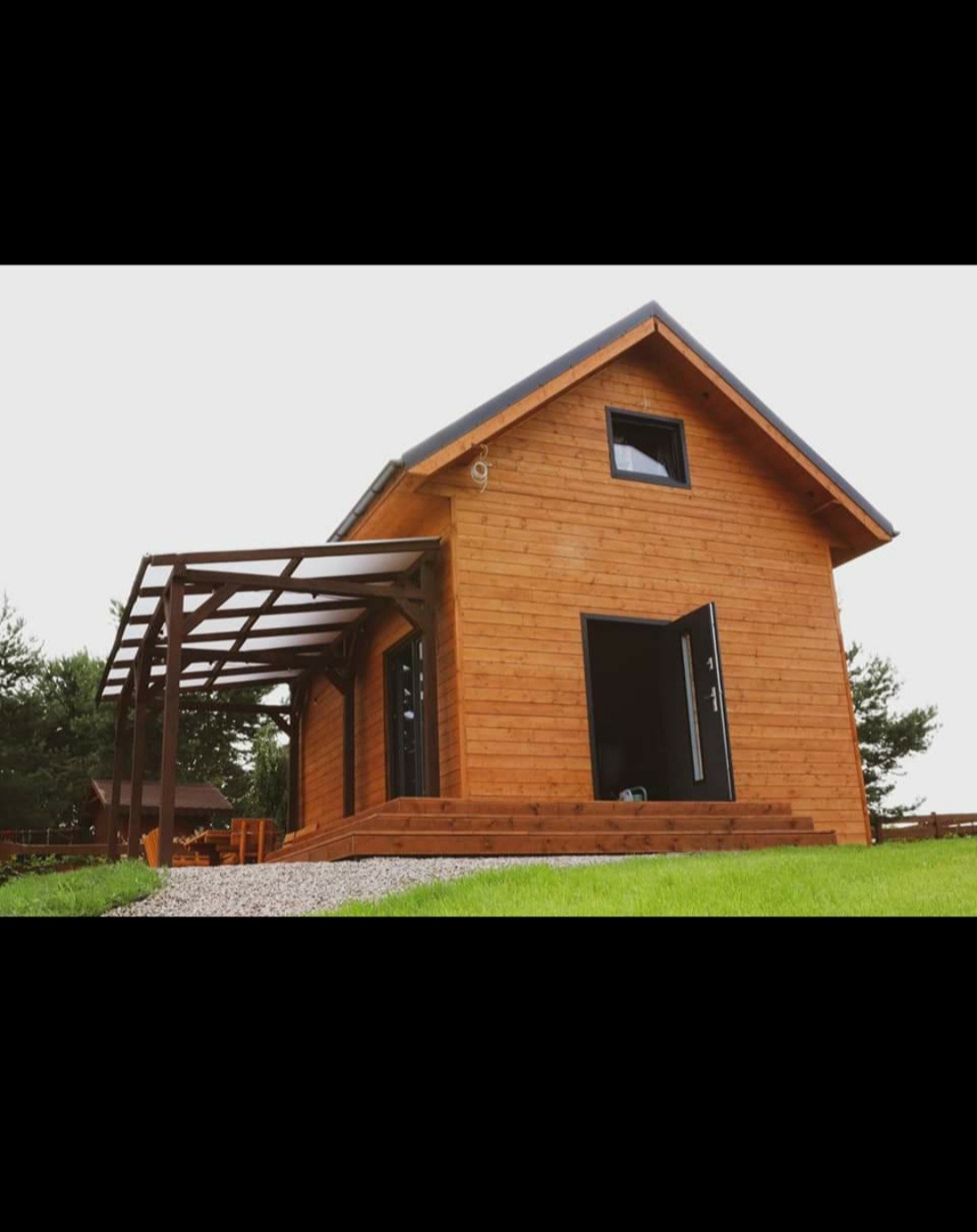 Drewniany domek z otwartymi drzwiami wejściowymi i zadaszoną pergolą, widok z poziomu trawnika.