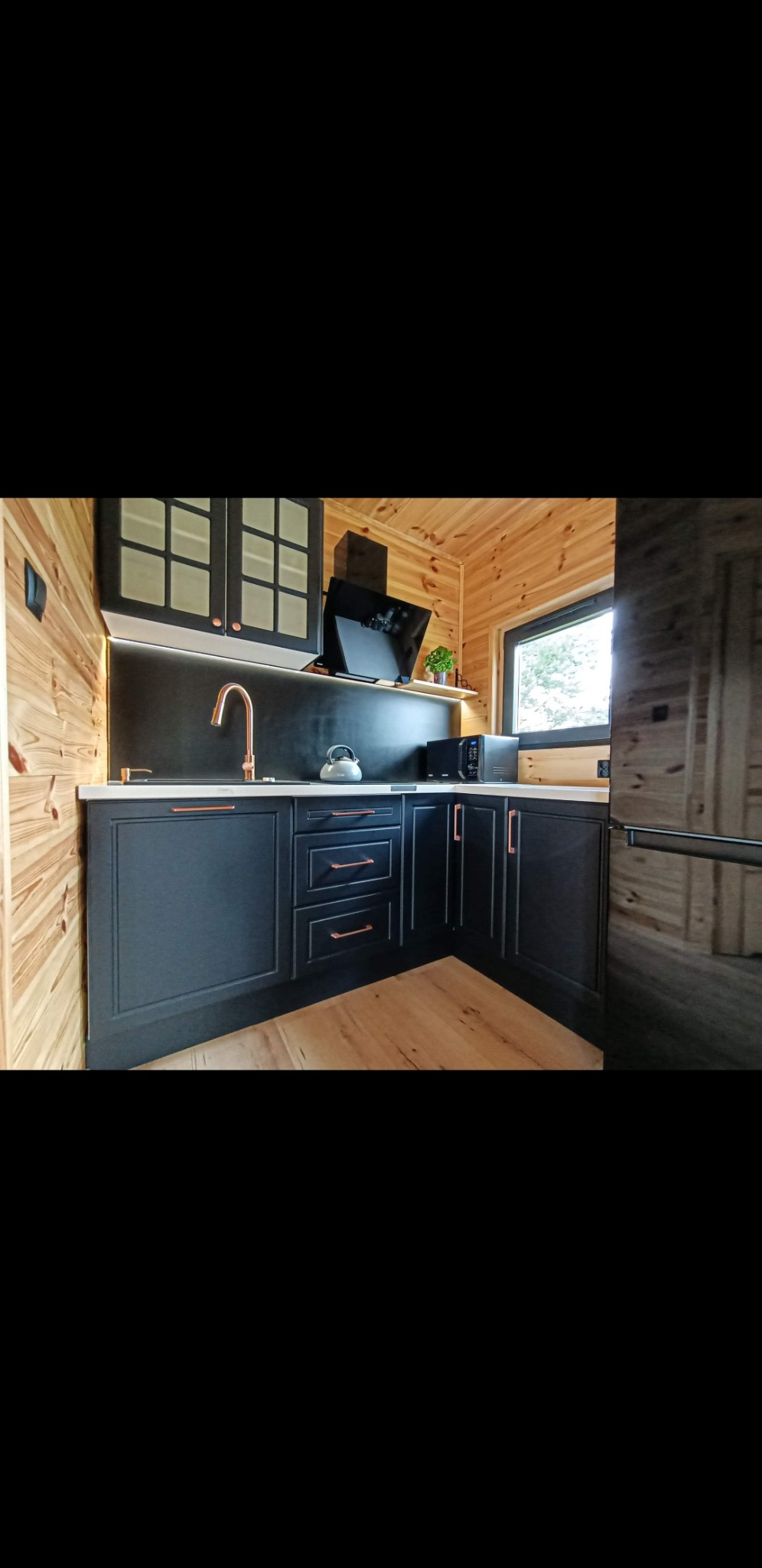 Kąt ujęcia na czarną, drewnianą zabudowę kuchenną z miedzianymi uchwytami, na tle ścian z jasnego drewna i podświetleniem LED pod szafkami wiszącymi.