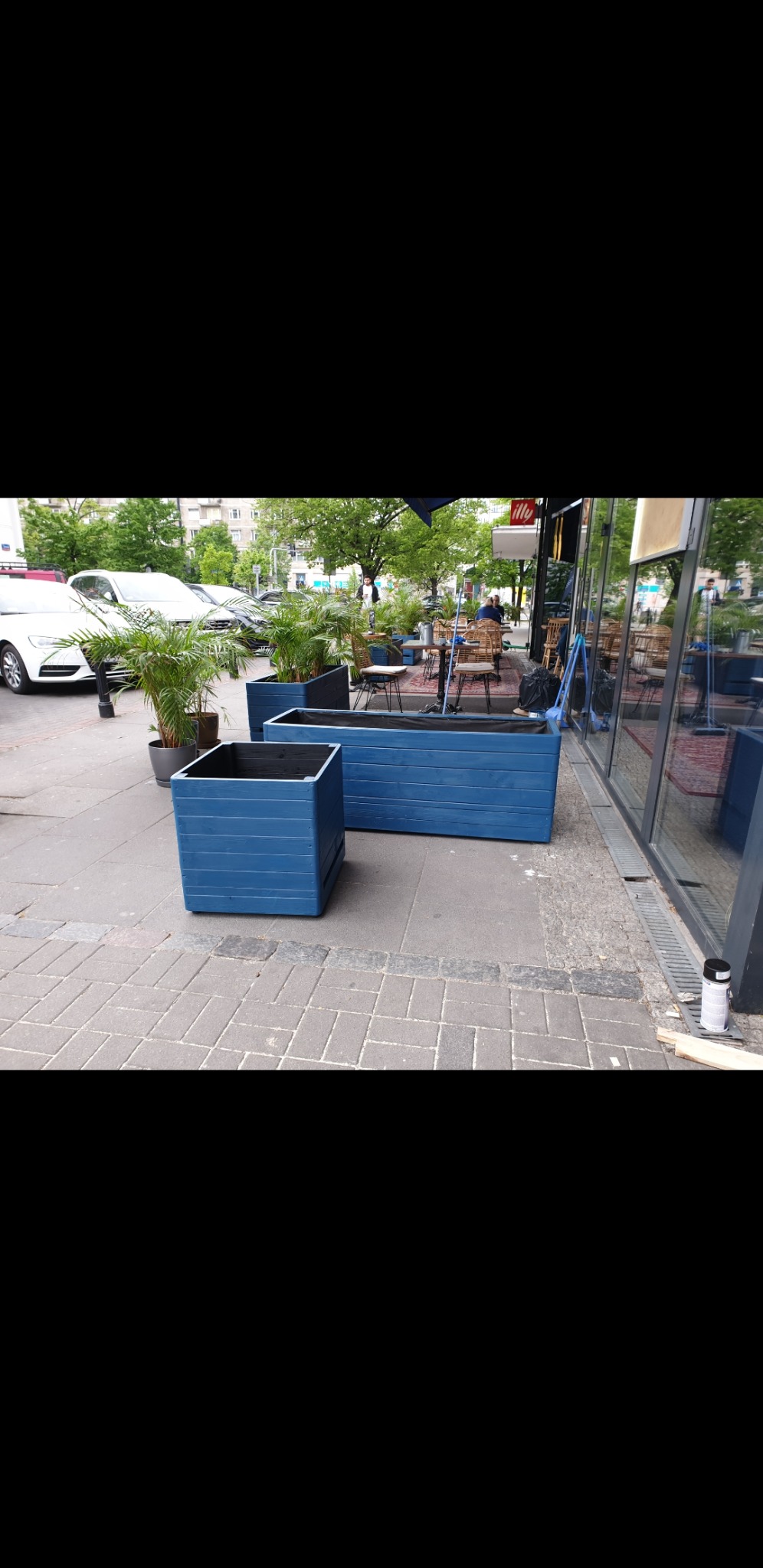 Niebieskie, drewniane donice na zewnątrz lokalu gastronomicznego, z widocznymi roślinami w tle i fragmentem chodnika z kostki.