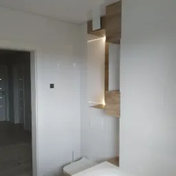 Nowoczesna, minimalistyczna łazienka z białymi, błyszczącymi płytkami, podświetlanymi drewnianymi półkami i widoczną częścią wanny.