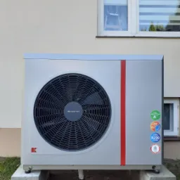 Zewnętrzna jednostka pompy ciepła marki Kolton z czerwoną pionową linią, zamontowana na betonowych blokach przed budynkiem z dwoma oknami.