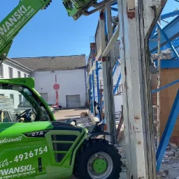 Zielony wózek widłowy teleskopowy demontuje metalową konstrukcję budynku, widoczny pracownik w okularach ochronnych na górze konstrukcji.