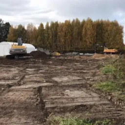 Teren budowy z dwoma koparkami, widoczne ślady wykopów i przygotowania pod fundamenty, w tle biały namiot i drzewa.