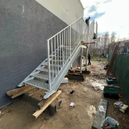 Zewnętrzne metalowe schody ewakuacyjne z kratownicowymi stopniami, zamontowane przy szarej ścianie budynku, widoczne narzędzia i materiały budowlane na placu budowy.