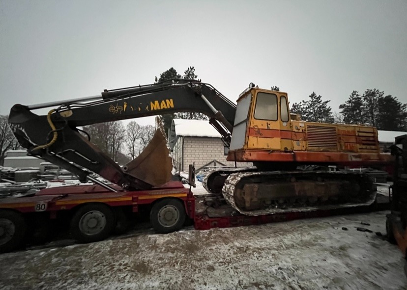 Żółto-pomarańczowa koparka marki MAN, załadowana na czerwoną niskopodwoziową lawetę, zimowy krajobraz w tle.