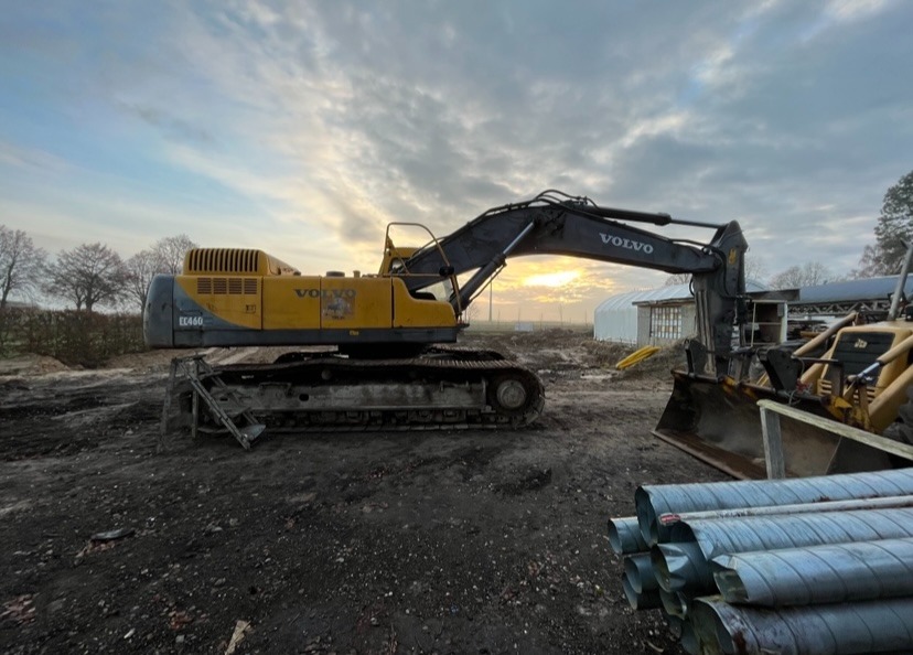Żółta koparka Volvo EC460 na placu budowy o zmierzchu, z rurami kanalizacyjnymi na pierwszym planie i widocznym budynkiem w tle.