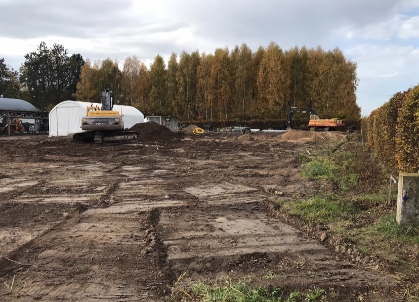 Teren budowy z dwoma koparkami, widoczne ślady wykopów i przygotowania pod fundamenty, w tle biały namiot i drzewa.