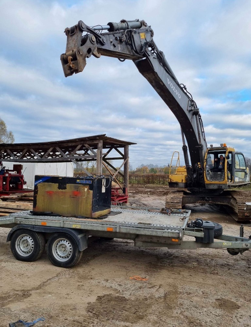 Ciężka koparka Volvo z hydraulicznymi nożycami do kruszenia betonu, przygotowana do transportu na niskopodwoziowej przyczepie, na placu budowy z drewnianą wiatą w tle.
