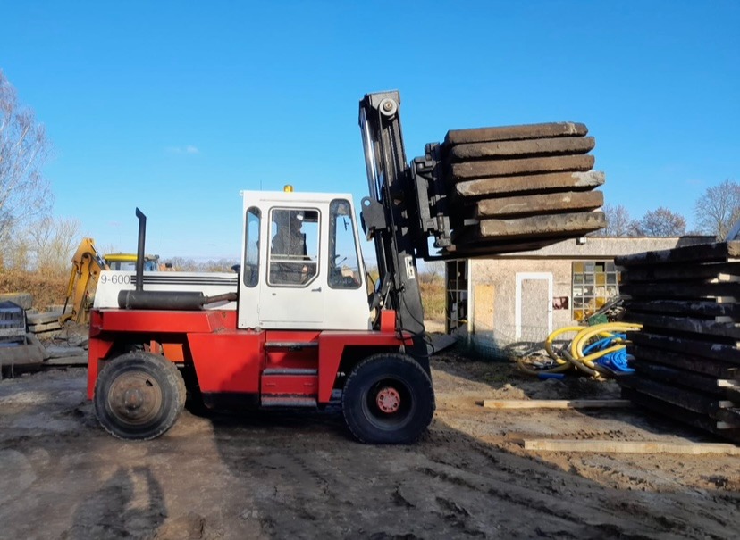 Czerwono-biały wózek widłowy transportuje ułożone płyty chodnikowe na placu budowy, w tle żółta koparka i budynek gospodarczy.