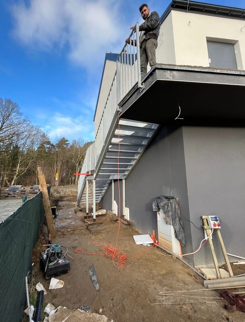 Montaż zewnętrznych schodów stalowych do budynku z płaskim dachem, widok z dołu, pracownik na górze, teren budowy z narzędziami i kablami.