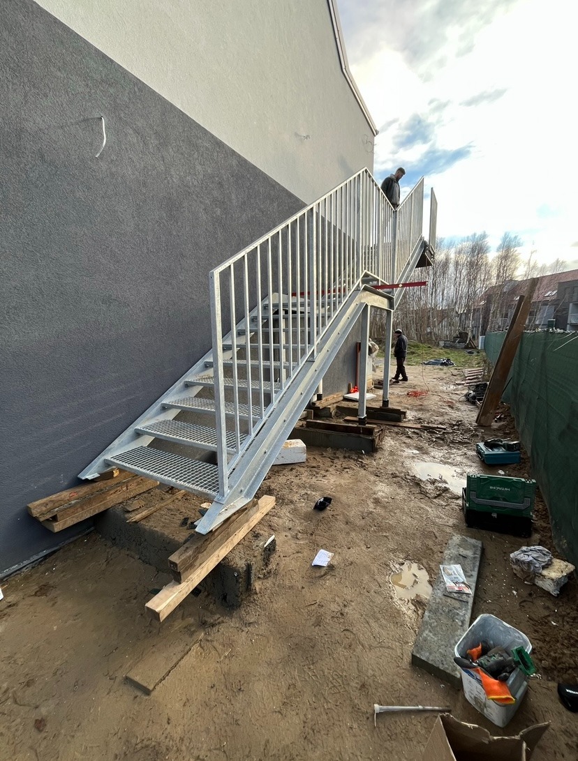 Zewnętrzne metalowe schody ewakuacyjne z kratownicowymi stopniami, zamontowane przy szarej ścianie budynku, widoczne narzędzia i materiały budowlane na placu budowy.