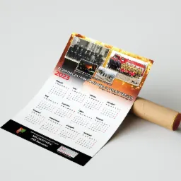 Kalendarz ścienny 2023 z motywem Ochotniczej Straży Pożarnej w Stanowicach, zawierający zdjęcia archiwalne i współczesne strażaków oraz wozów strażackich, zwinięty i oparty o tubę.