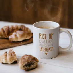 Kawa parująca w białym kubku z napisem 'Rise up & attack the day with enthusiasm' stoi na białym drewnianym stole obok croissantów na desce i luzem.