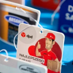 Kolekcja różnorodnych, kolorowych zawieszek zapachowych z nadrukami, w tym DPD z kurierem, logo firm i hasła reklamowe na błękitnym tle.
