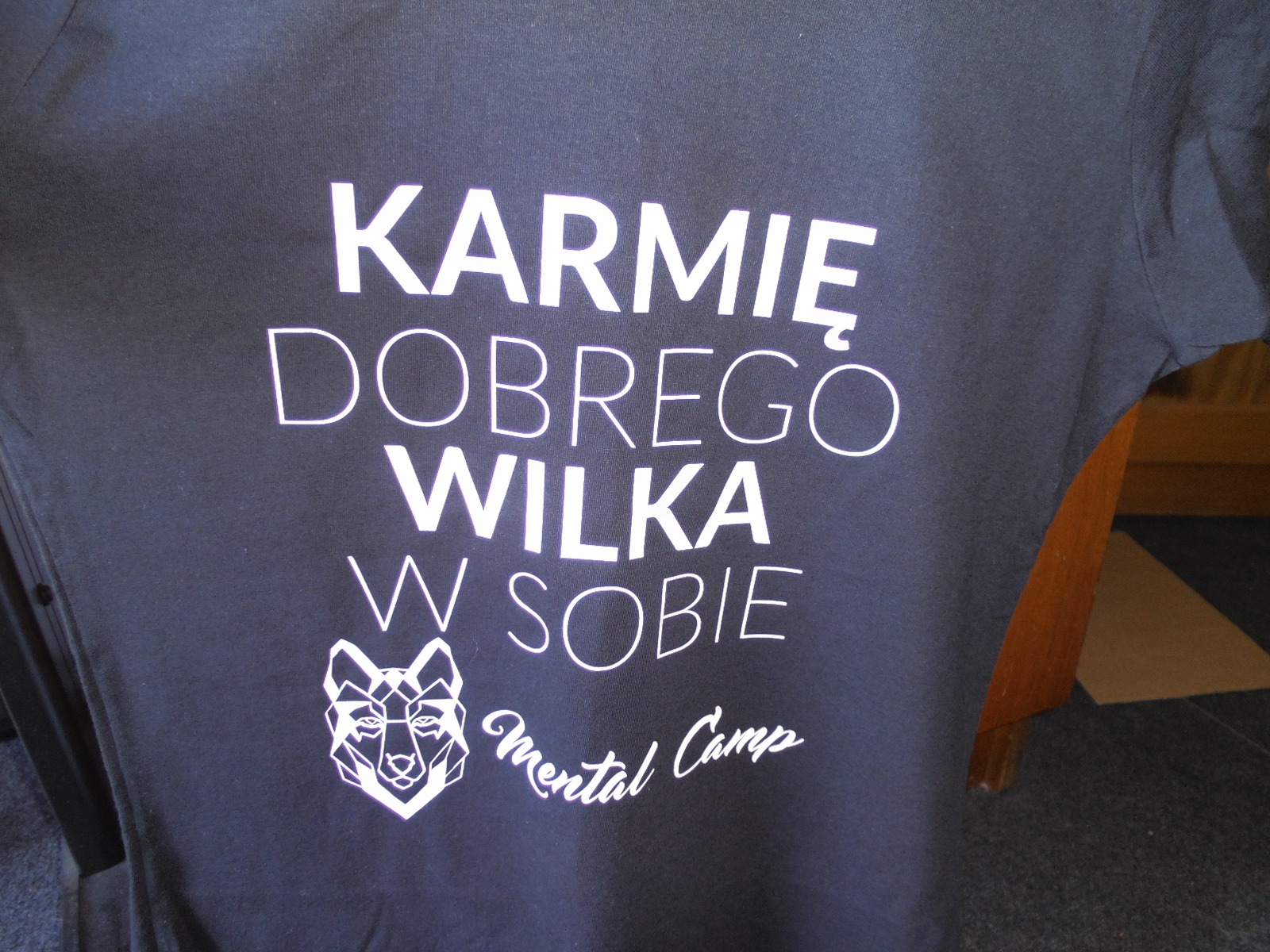 Czarna koszulka z białym nadrukiem: 'Karmię dobrego wilka w sobie' oraz grafika głowy wilka i napis 'Mental Camp'.