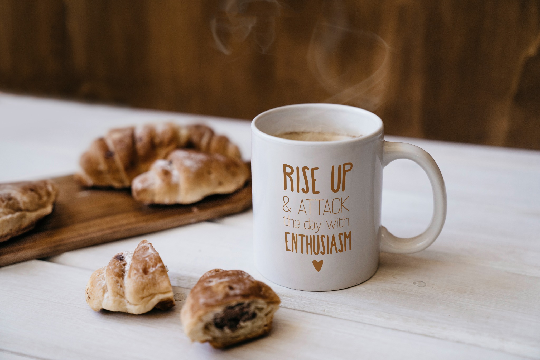 Kawa parująca w białym kubku z napisem 'Rise up & attack the day with enthusiasm' stoi na białym drewnianym stole obok croissantów na desce i luzem.