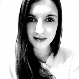 Czarno-biały portret młodej kobiety z długimi, ciemnymi włosami, uśmiechającej się delikatnie w kierunku kamery, z oświetleniem podkreślającym rysy twarzy.