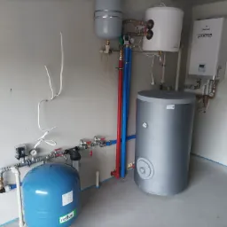Kompletna instalacja grzewcza z kotłem gazowym Galmet Prima, zbiornikiem akumulacyjnym i naczyniem przeponowym Reflex, rury w kolorach czerwonym i niebieskim, zamontowana w surowym pomieszczeniu.
