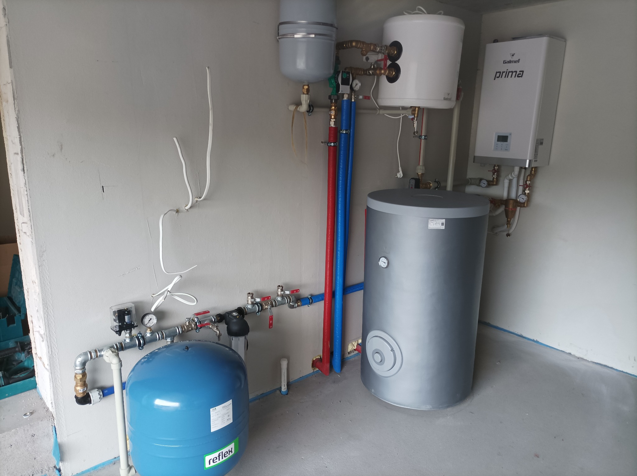 Kompletna instalacja grzewcza z kotłem gazowym Galmet Prima, zbiornikiem akumulacyjnym i naczyniem przeponowym Reflex, rury w kolorach czerwonym i niebieskim, zamontowana w surowym pomieszczeniu.