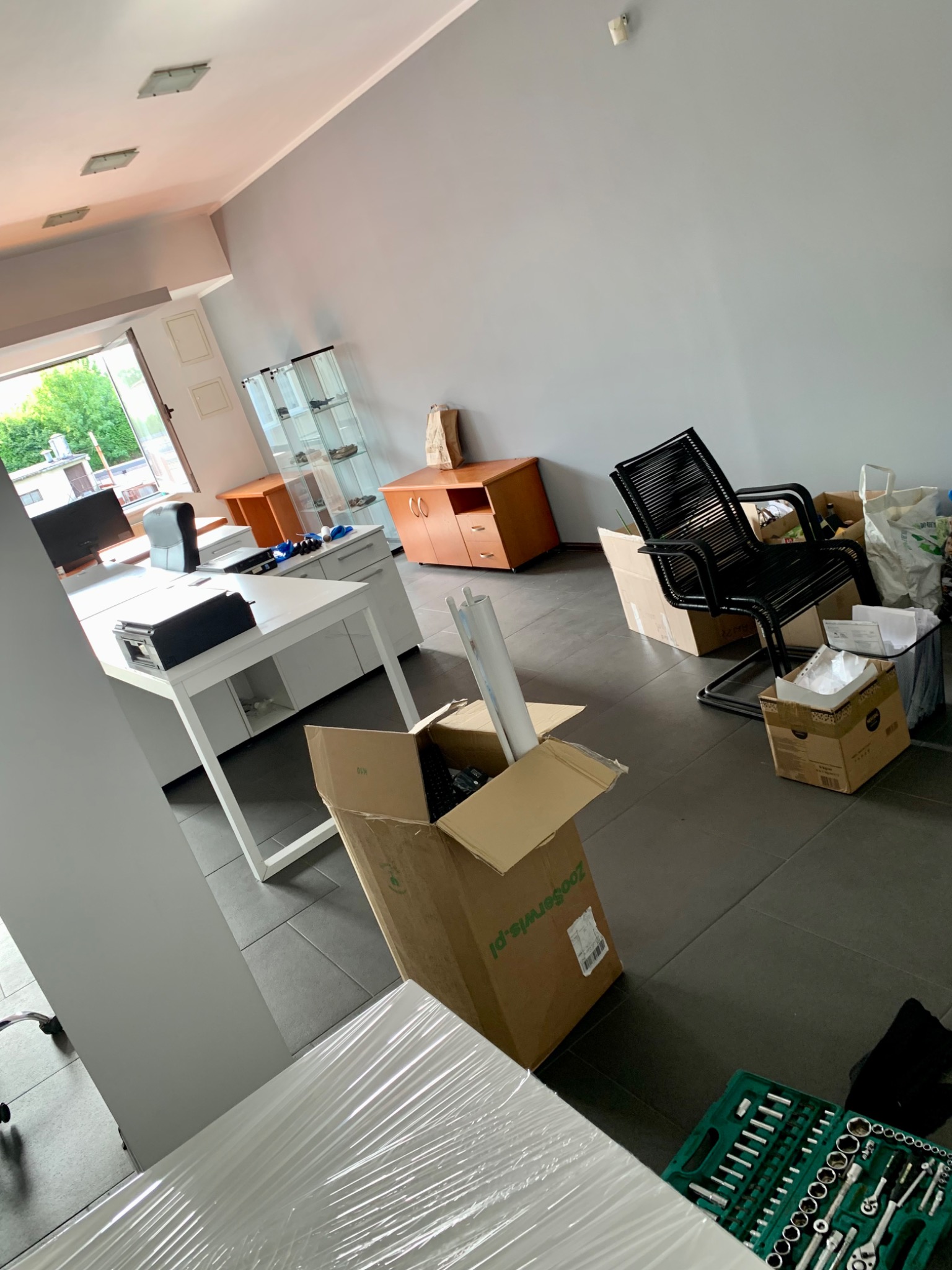 Biuro w trakcie przeprowadzki: zapakowane biurka owinięte folią stretch, kartony z zawartością, krzesło biurowe i zestaw narzędzi na podłodze w nowym lokalu.