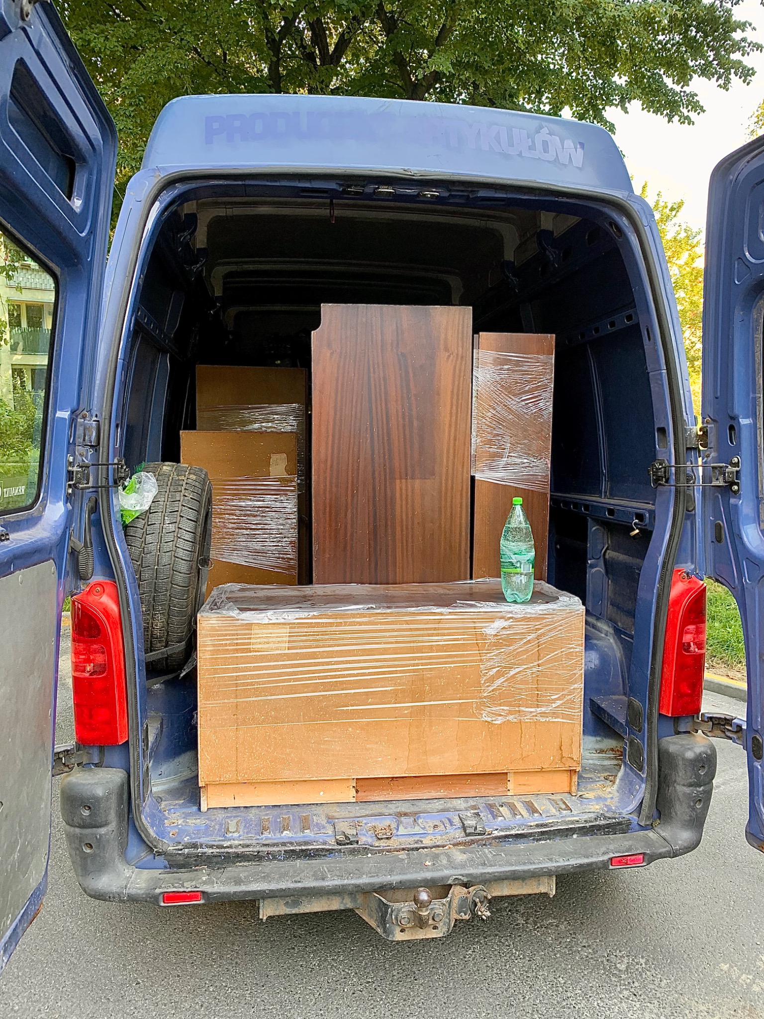 Wnętrze niebieskiego busa z otwartymi drzwiami załadowane meblami zabezpieczonymi folią stretch, widoczna opona i butelka wody.