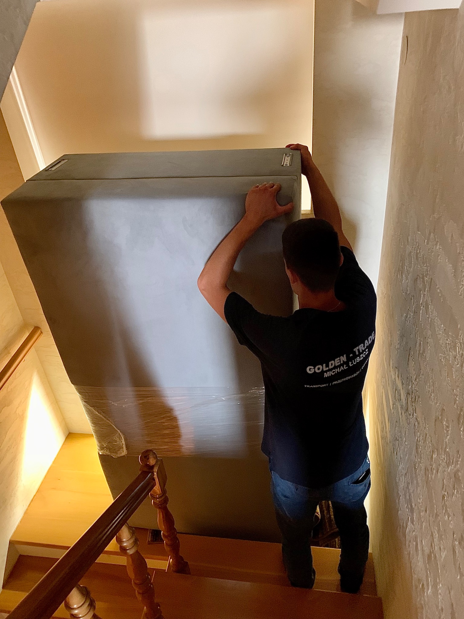 Pracownik firmy przeprowadzkowej w czarnej koszulce z logo 'GOLDEN TRADE' wnosi po schodach duży, owinięty folią mebel.