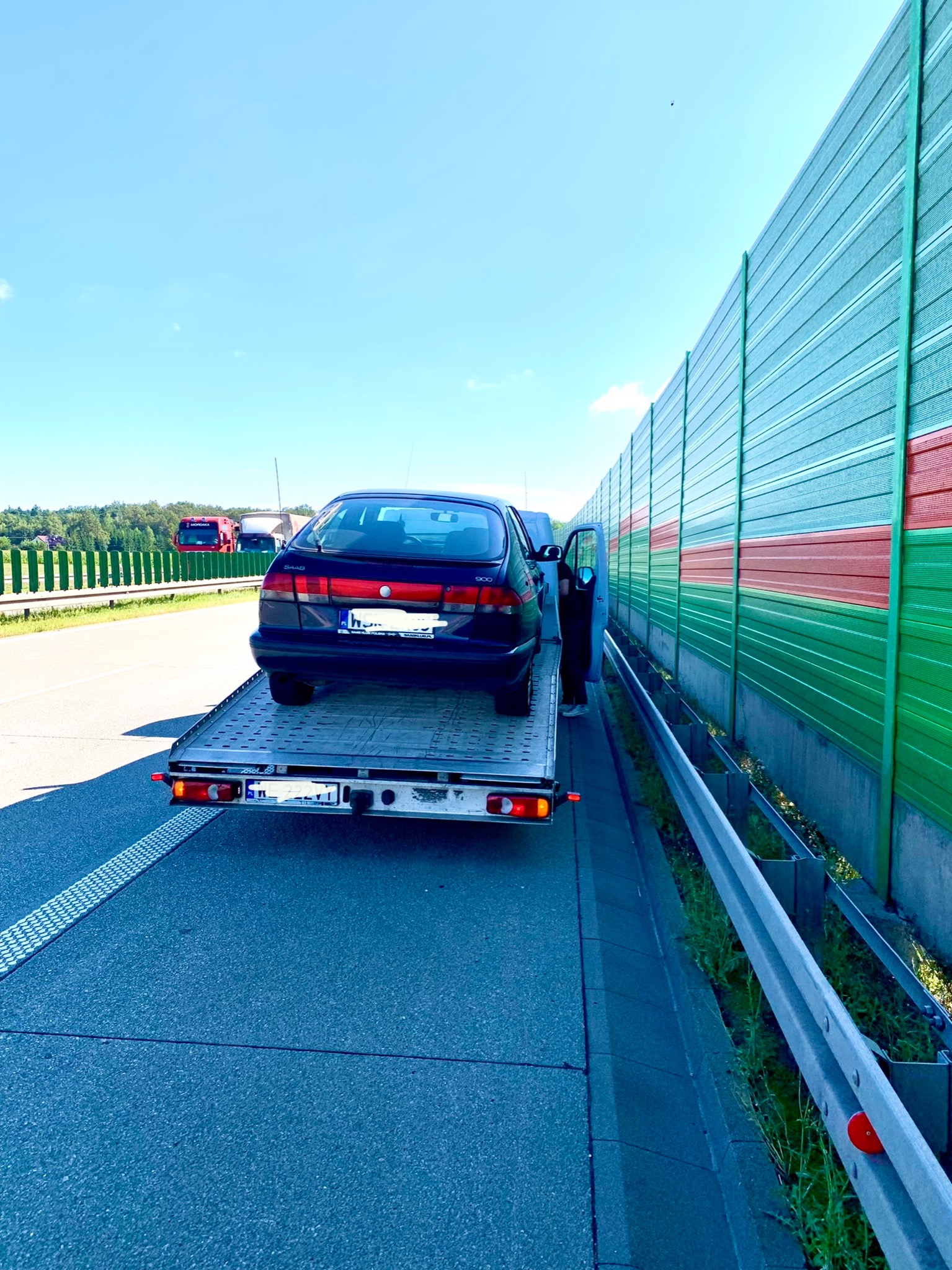 Czarny samochód Saab 900 na platformie lawety, zaparkowany na poboczu autostrady z otwartymi drzwiami pasażera, widok z tyłu, w tle bariera dźwiękochłonna i inne pojazdy.