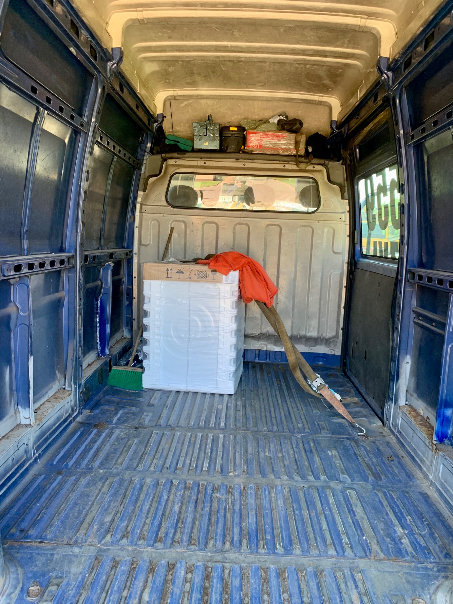 Wnętrze niebieskiego busa transportowego z zabezpieczonym ładunkiem: karton owinięty folią, przytrzymywany pasem transportowym i częściowo przykryty pomarańczową tkaniną, dodatkowo widoczna miotła