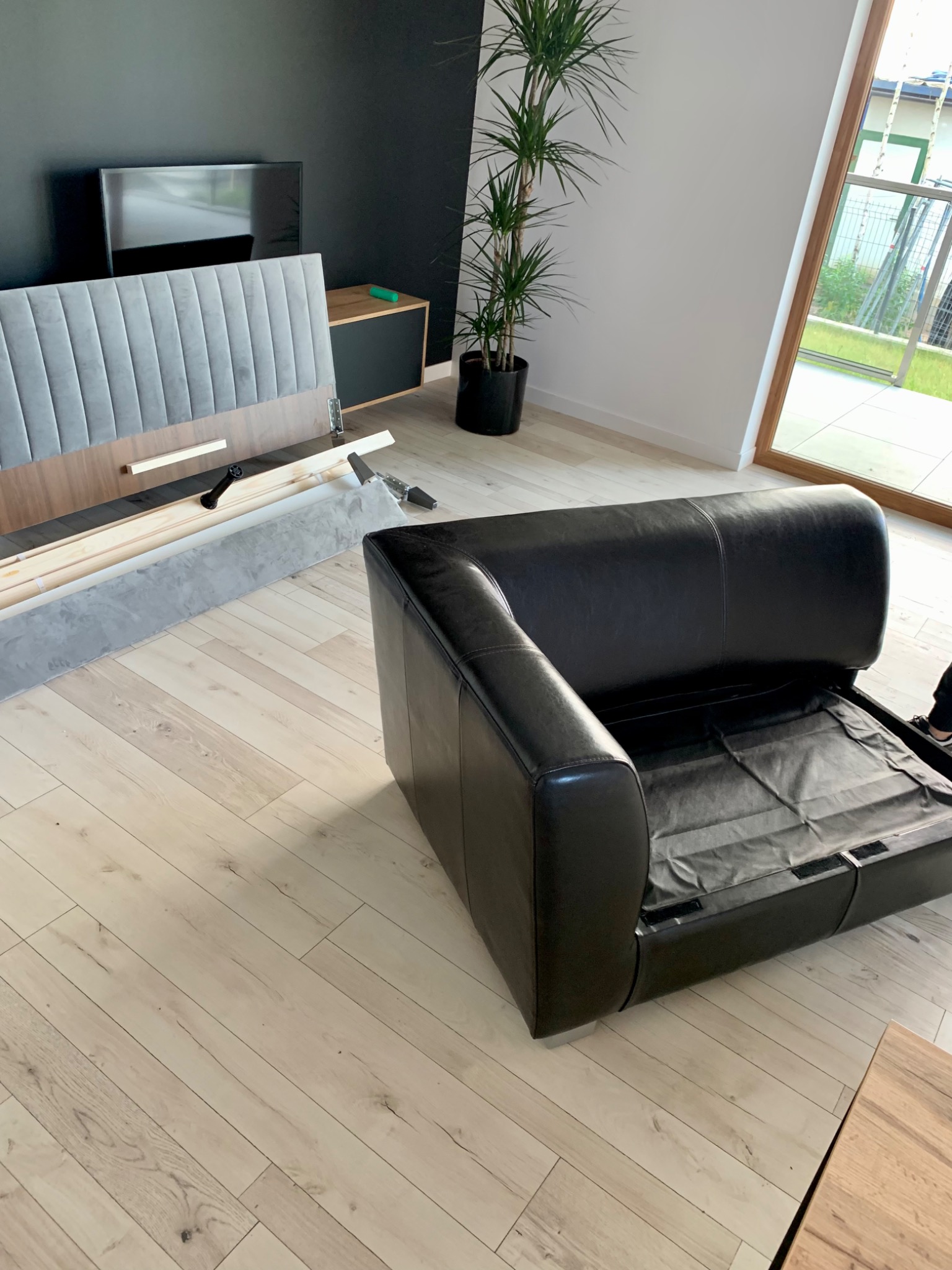 Rozłożone elementy tapicerowanego łóżka z szarym wezgłowiem i czarny skórzany fotel w trakcie montażu w jasnym pokoju z drewnianą podłogą.