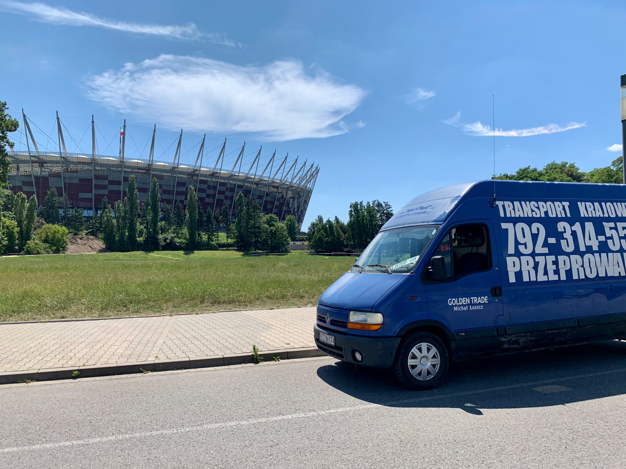 Niebieski bus z napisem 'TRANSPORT KRAJOWY, 792-314-555, PRZEPROWADZKI' na tle Stadionu Narodowego w Warszawie, widoczny fragment chodnika i jezdni.